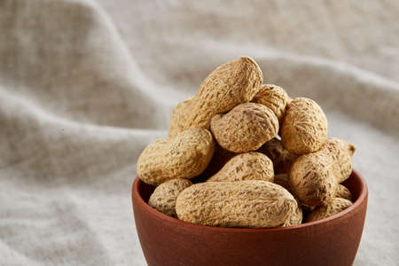 Composition of raw unpeeled peanuts in clay ceramic bowl on light grey cotton tablecloth or napkin,...