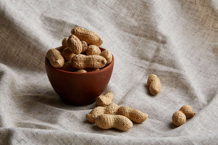 Composition of raw unpeeled peanuts in clay ceramic bowl on light grey cotton tablecloth or napkin,...
