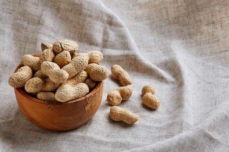 Composition of raw unpeeled peanuts in wooden bowl on light grey cotton tablecloth or napkin, some copy space, closeup, selective focus. Healthy lifestyle concept.の写真素材