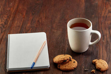 A white inside and brown outside porcelain cup of tea with tasty chocolate chips cookies, empty workbook and long yellow pencil with blue top on a dark wooden vintage background, top view.の写真素材
