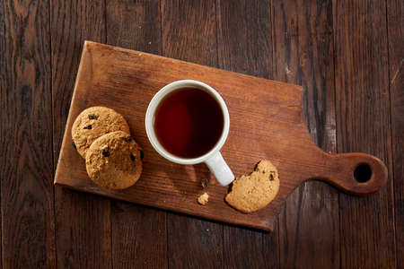 A white inside and brown outside porcelain cup of tea with tasty chocolate chips cookies, empty workbook and long yellow pencil with blue top on a dark wooden vintage background, top view.の写真素材