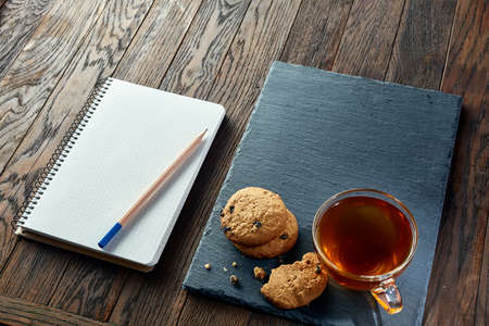 A white inside and brown outside porcelain cup of tea with tasty chocolate chips cookies, empty workbook and long yellow pencil with blue top on a dark wooden vintage background, top view.の写真素材