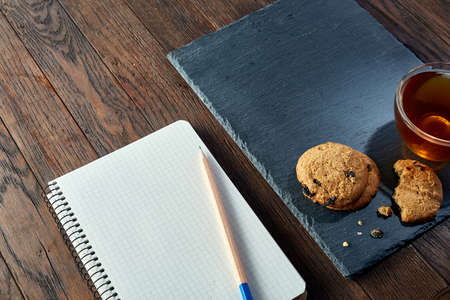 A white inside and brown outside porcelain cup of tea with tasty chocolate chips cookies, empty workbook and long yellow pencil with blue top on a dark wooden vintage background, top view.の写真素材