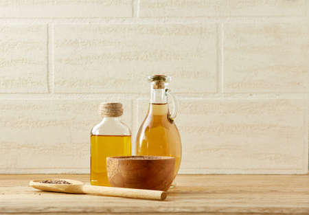 Composition of two transparent glass oil jars, wooden bowl with flax seeds and a scoop on wooden table over a light background, selective focus. Kitchen utensils and ingredients on wooden table. Healthy food concept.の写真素材