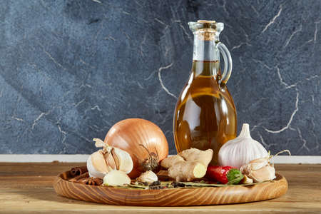 Conceptual composition of different spices and transparent glass jar of oil on a wooden tray on a rustic table: onion, garlic, cloves, chilly, ginger, anise, cinnamon, peppercorn, bay leaves, top view. Picturesque still life.の写真素材