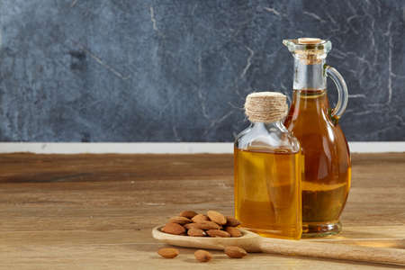 Composition of aromatic oil in a glass jar and bottle with unpeeled almond in a scoop on wooden table over a dark marble background, close-up. Nutritious therapeutic food for healthy lifestyle.の写真素材