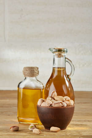 Composition of aromatic oil in a glass jar and bottle with unpeeled pistachios in bowl on wooden table over a light background, close-up. Nutritious therapeutic food for healthy lifestyle.の写真素材