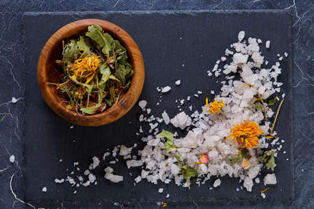 Pile of salt crystals mixed with motley grass and wooden bowl on the dark background, close-up, top view, selective focus. Some copy space for your text or inscription. Abstract background. Spa and wellness setting. Spa decoration concept.の写真素材