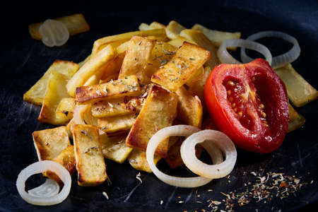 Delicious homemade spicy fries with onion and grilled tomato over dark background, selective focus