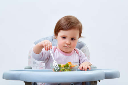 Adorable toddler girl is having fun while eating stewed vegetables on white backgroundの写真素材