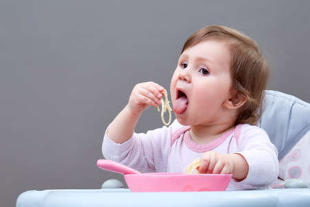 Adorable toddler girl is having fun while eating stewed vegetables on grey backgroundの写真素材