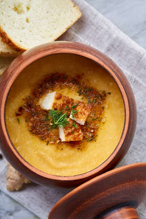 Clay pot of spicy creamy pumpkin soup garnished with dill, served with toasted baguette bread slices...