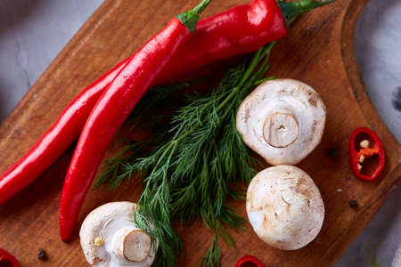 Top view of cutting board with red hot pepper, mushrooms, dill, parsley on wooden tabletop, selective focus, shallow depth of field. Culinary still life. Healthy eating. Seasonal countryside cuisine. Food concept.の写真素材