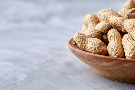 Unpeeled peanuts in wooden bowl over white textured background closeup, selective focus. Some copy...