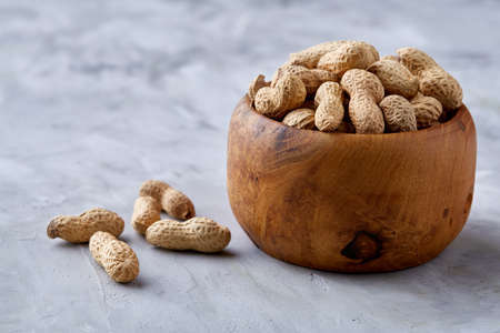 Unpeeled peanuts in wooden bowl with some nuts next to it over white textured background closeup,...