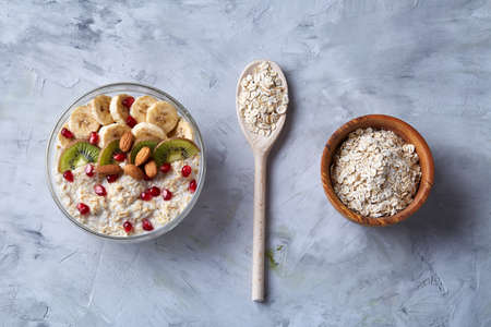 Diet breakfast oatmeal cereal with kiwi, banana, almond and pomergranate seeds and wooden bowl and spoon with oat flakes over white textured background, selective focus, close-up, top view. Healthy eating concept. Breakfast background.の写真素材