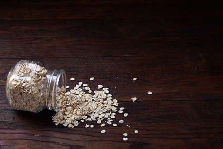 Overturned glass jar and wooden spoon with raw oatmeal on vintage wooden background, close-up, top view, selective focus. Healthy breakfast concept with uncooked oatmeal on a wooden background.の写真素材