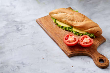Fresh and tasty submarine sandwich with cheese and vegetables with tomato half on cutting board over white textured background, selective focus, top view. Nutritious crusty snack. Yummy fast food. American cuisine. Food background.の写真素材