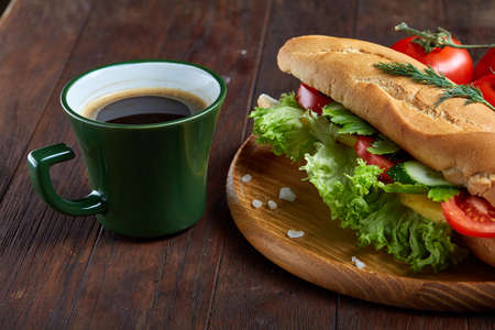 Fresh homemade sub sandwich with lettuce, tomatoes and cheese served with fresh tomatoes, cup of refreshing coffee on wooden plate over vintage wooden background, selective focus. Ready to eat tasty snack. American cuisine. Food concept.の写真素材