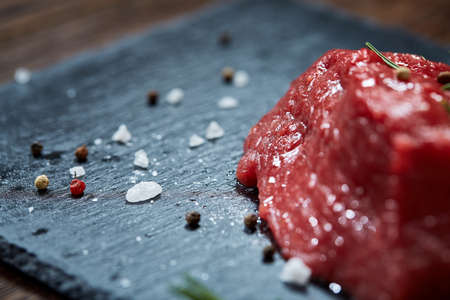 Raw meat beef steaks on black slate board with spices, garlic and rosemary over wooden background,...
