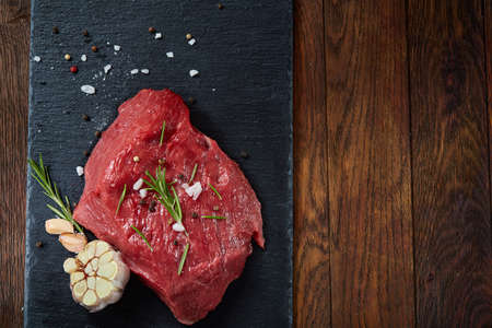 Raw meat beef steaks on black slate board with spices, garlic and rosemary over wooden background, copy space, selective focus, shallow depth of field. Gourmet beesteak. Organic protein. Nutritious butcher. Healthy food concept.の写真素材