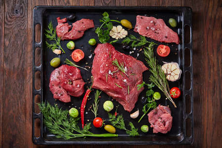 Flat lay of raw beefsteak with tomatoes, garlic, hot pepper, dill, parsley, rosemary and spicies on metal cooking tray, close-up, selective focus. Preparation for cooking. Hight quality barbeque. Gourmet beesteak. Organic protein. Food concept.の写真素材