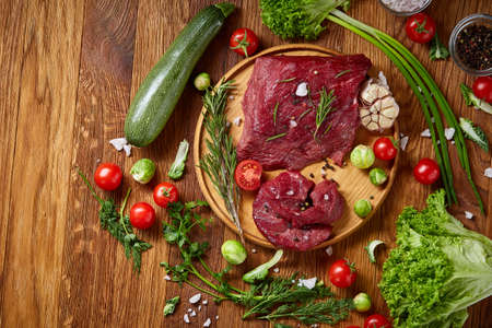 Still life of raw beef meat framed with tomatoes, garlic, brussels sprouts, dill, parsley, rosemary and spicies on wooden plate over white background, top view, selective focus. Preparation for cooking. Organic protein. Food concept.の写真素材