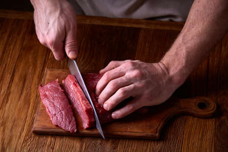 Preparation for dinner. Cooking, processing of meat. Strong professional man's hands cutting raw beefsteak, selective focus, close-up. Self-confident chef on his work. Studio shot. Healthy eating dieting. Food background.の写真素材