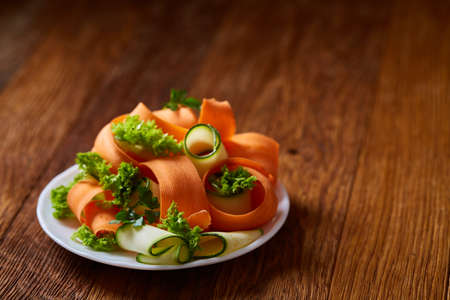 Artistically served vegetable salad with carrot, cucumber, letucce on white plate over wooden background, selective focus. Some copy space. Culinary masterpiece. Restaurant serving. Gourmet low fat dish. Healthy eating dieting.の写真素材
