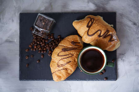 Coffee cup, overturned jar with coffee beans, croissants on stone board over white textured background, selective focus, close-up. Studio shot. Delicious morning beverage. Time for calm and relax. Tasty dessert. Food concept.の写真素材