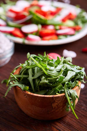 Fresh ruccola in a wooden bowl on wooden table over vegetable background, close-up, selective focus, shallow depth of field, vertical. Fresh arugula leaves. Organic vitamin source. Tasty salad ingredient. Healthy food concept.の写真素材
