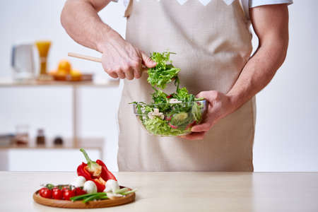 Man cooking at kitchen making healthy vegetable salad, close-up, selective focus. Variety of vegetables on wooden plate on white table in front of the chef. Skilful chef in an apron mixing fresh sald ingredients. Cooking and home conceptの写真素材