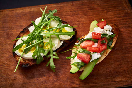 Grilled toast brisket with tomato, avocado and ruccola on wooden cutting board over dark background, top view, close-up, selective focus. Perfect vegetarian sandwich. Tasty snack for Lent. Food concept.の写真素材