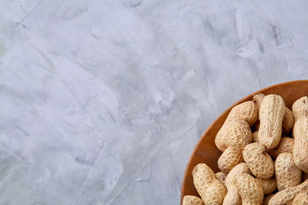 Unpeeled peanuts in wooden bowl over white textured background closeup, selective focus. Healthful...