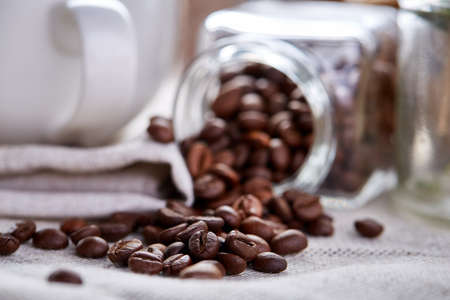 Side view of natural roasted coffee beans get out of overturned glass jar on table covered with white homespun tablecloth, selective focus. Caffeinated composition. Stimulating beverage. Coffee beans background. Studio shot. Natural brewed drink.の写真素材