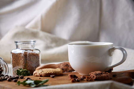 Breakfast background with porcelain mug of fresh black coffee, homemade oatmeal cookies on cutting board, glass jar of grind coffee on white cotton homespun tablecloth, close-up, selective focus. Studio shot. Rustic composition. Food concept.の写真素材
