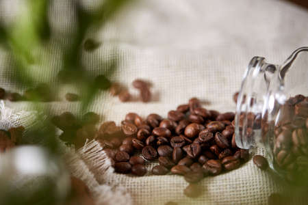 Side view of natural roasted coffee beans get out of overturned glass jar on homespun tablecloth, selective focus, back focus. Caffeinated composition. Stimulating beverage. Coffee beans background. Studio shot. Natural brewed drink.の写真素材