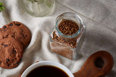 Breakfast background with porcelain mug of fresh black coffee, homemade oatmeal cookies on cutting board, glass jar of grind coffee on white cotton homespun tablecloth, close-up, selective focus. Studio shot. Rustic composition. Food concept.の写真素材