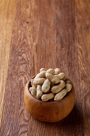 Unpeeled peanuts in wooden bowl over rustic wooden background closeup, selective focus. Some copy...
