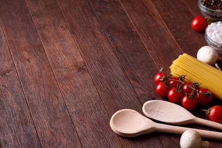 Uncooked pasta, tomatoes, mushrooms and two wooden spoon on rustic wooden background, top view,...