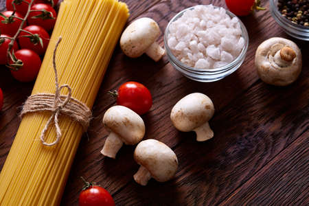 Uncooked pasta, tomatoes, mushrooms and two wooden spoon on rustic wooden background, top view, close-up, selective focus. Preparation for cooking. Italian cuisine. Ingredients for perfect mediterranian meal. Food concept.の写真素材