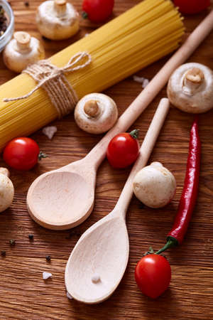 Uncooked pasta, tomatoes, mushrooms and two wooden spoon on rustic wooden background, top view, close-up, selective focus. Preparation for cooking. Italian cuisine. Ingredients for perfect mediterranian meal. Food concept.の写真素材
