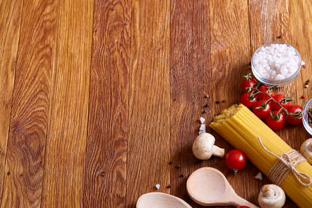Uncooked pasta, tomatoes, mushrooms and two wooden spoon on rustic wooden background, top view, close-up, selective focus. Preparation for cooking. Italian cuisine. Ingredients for perfect mediterranian meal. Food concept.の写真素材