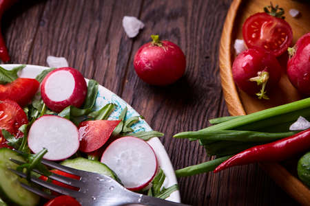 Creative fresh vegetable salad with ruccola, cucumber, tomatoes and raddish on white plate over wooden background, close-up, selective focus. Morning atmospheric lighting, fashionable trendy spot. Preparation for design creative menu. Food conceptの写真素材