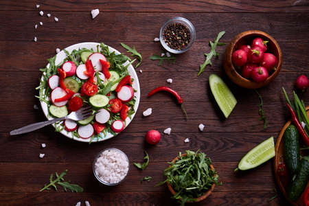 Fresh red radish in wooden bowl among plates with vegetables, ruccola and spicies on rustic wooden background, close-up, selective focus, shallow depth of field. Freshly gathered seasoning. Delicious appetizer. Summer garnish. Healthy food concept.の写真素材