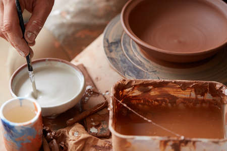 A potter in a plaid shirt and apron prepare paints to dye a clay plate in white in workshop, close-up, selective focus. Some tools, clay samples and paint jars in the background. Aristic manufacturing. Creative hobby. Art and crafts concept.の写真素材