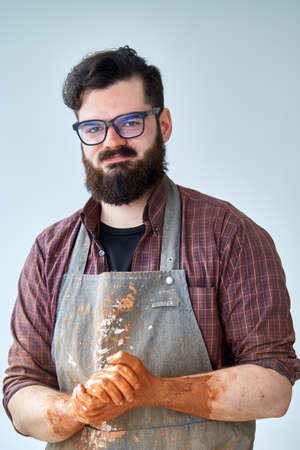 Portrait of handsome young bearded potter posing in studio, close-up. Artistic craftsman wearing plaid shirt and dirty apron holding his hands covered with fresh clay in front of him. Guy is wearing glasses and stylish haircut. Cheerful ceramist.の写真素材