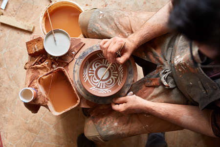 A potter in a plaid shirt and apron paints a clay plate in white in workshop, top view, close-up, selective focus. There are some tools, clay samples and paint jars in the background. Aristic manufacturing. Creative hobby. Art and crafts concept.の写真素材