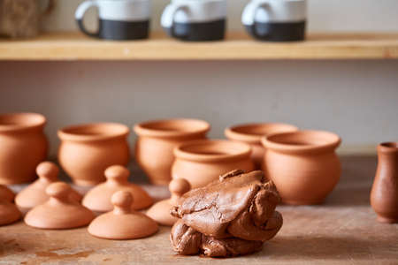 Ceramic dishware on worktop and shelves with piece of clay in front of in pottery workshop,...
