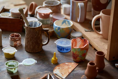 Close-up of various paint mugs and brushes in holder on worktop over wooden shelves with pottery, selective focus, side view. Preparing for decoration. Creative lay out. Handwork lifestyle. Handicraft conceptの写真素材